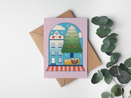 Colorful holiday greeting card featuring a snowy winter scene with a house, tree, and snowflakes. The card has a whimsical, illustrated style.