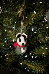 A plush Christmas ornament in the shape of a badger, with a red scarf and hanging from a Christmas tree.