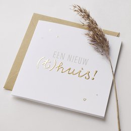 A greeting card with the text "Een nieuw huis!" (A new house!) in gold foil lettering on a white background, accompanied by a small dried plant stem. The card is presented with a matching envelope.