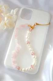 A white phone case with a delicate rose quartz and freshwater pearl lanyard. The lanyard features a gold-tone chain and clasp, with pink and white pearls strung along its length.