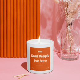 Tall, round orange scented candle with the text "Cool People live here" printed on the label. The candle is surrounded by pink and orange decor elements, creating a cozy and stylish atmosphere.