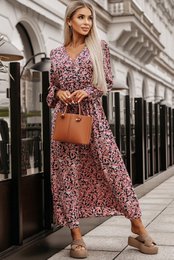 A long, floral print dress with a cinched waist and long sleeves. The dress features a v-neckline and a flowing, ankle-length skirt. The model is carrying a brown leather handbag and wearing beige sandals.