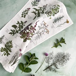 Organic tea towel featuring a botanical print with various herbs and plants. The towel has a white background with green, purple, and brown illustrations of different herbs and leaves.