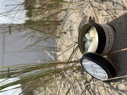 Wax melts in a round container with a label that says "L'eau de Mer" and features a sea salt and driftwood scent. The wax melts are surrounded by green moss-covered rocks, creating a natural and earthy aesthetic.