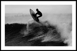 Surfer catching waves on a black and white poster. The poster features a surfer riding a large wave, creating a dramatic and dynamic image. The poster is available in various sizes, including framed and canvas options, making it suitable for home or office decor.