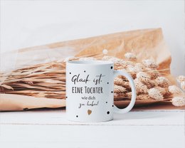 A white ceramic mug with the text "Endlich Schulkind" (Finally a school child) and colorful polka dot patterns. The mug is being held by a person's hands against a striped background.
