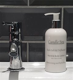 A tall, round white bottle of Candle Spa Hand & Body Lotion in the Rock Salt & Driftwood scent. The bottle has a pump dispenser and is placed on a dark surface next to a metal decorative item.