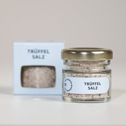Truffle salt in a small glass jar with a white label. The salt appears to be a light gray color with small black specks, indicating the presence of truffles. The product is packaged in a simple, minimalist style.