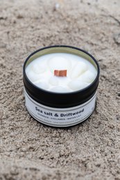 Scented candle in a travel tin, featuring a sea salt and driftwood fragrance. The candle has a white wax and is presented in a black tin container.