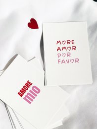 Red heart-shaped sticker, white greeting card with "MORE AMOR POR FAVOR" text, black pen.