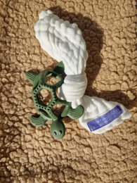 A green and white turtle-shaped teething ring with a soft, textured surface and a small green turtle figurine attached.