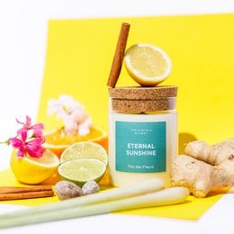A tall, round, yellow scented candle with a cork lid and a lemon slice on top, surrounded by fresh ginger, lemon slices, and pink flowers on a yellow background.