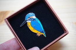 Colorful brooch featuring a detailed image of a vibrant blue and orange kingfisher bird. The brooch has a shiny, textured surface and is presented in a wooden display box.