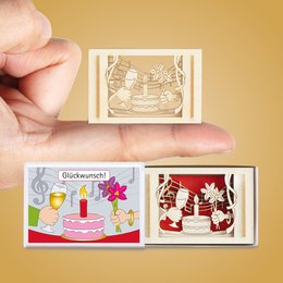 A small, decorative gift box featuring a silhouette design with various celebratory elements such as a cake, balloons, and confetti. The box is available in multiple languages, including German, English, French, and Dutch.