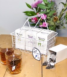 White wire serving basket with compartments for tea accessories, including tea bags, tea infuser, and tea cup.