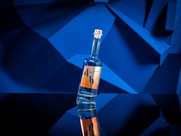 A bottle of The Drunken Horse Gin, a clear glass bottle with a blue and orange label, standing on a reflective surface against a dark blue background.