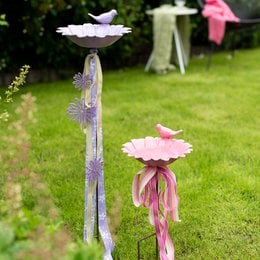 Decorative garden stakes with flower-shaped bird baths in cream, light orange, pink, and lavender colors. The stakes feature a rustic, weathered appearance and are suitable for use in outdoor garden settings.
