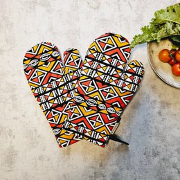 A pair of handmade oven gloves featuring a vibrant, geometric "Mudcloth" or Bogolan print inspired by traditional African textiles. The gloves have a bold, colorful pattern with a mix of triangles, rectangles, and other shapes in red, yellow, black, and white.