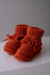 Rust red hand-knitted merino wool baby shoes with laces and a cozy, textured design.