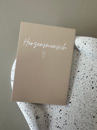 A small greeting card with the text "Herzensmensch" (German for "person of the heart") printed in white on a beige background.