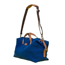 A blue canvas weekender bag with leather accents and handles. The bag has a structured shape and a zipper closure.