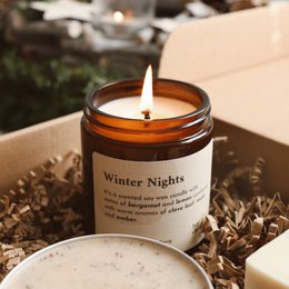 A round, tall, amber-colored glass candle jar with a white label that reads "Winter Nights". The candle is lit, casting a warm glow. The candle is surrounded by pine cones and other winter decor, suggesting a cozy, winter-themed scented candle.