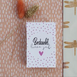 A small white card with the handwritten text "Bedankt!" (Thank you) and a small heart. The card is decorated with a feather and placed on a patterned background.