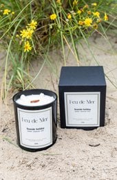 A tall, round, white scented candle with a floral design, set in a black container and surrounded by natural foliage.