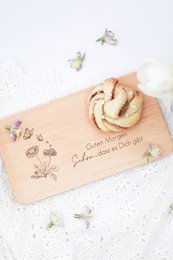  A wooden breakfast board with the text "Good morning. It's great that you're there!" Decorated with pine branches, pine cones, and various flowers.