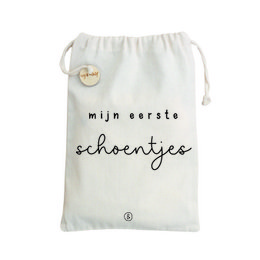 A white canvas storage bag for a child's first shoes, featuring the text "mijn eerste schoentjes" (my first shoes) in black lettering. The bag has a drawstring closure and a small gold-colored charm.