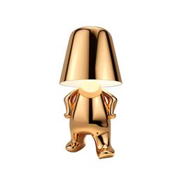  Gold metallic table lamp with decorative character base, modern sculptural design, shiny finish, unique artistic lighting fixture.