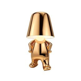 Gold metallic table lamp with decorative character base, modern sculptural design, shiny finish, unique artistic lighting fixture.
