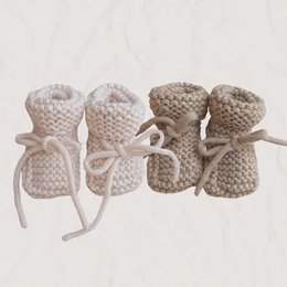 Knitted baby booties in ecru and beige colors, featuring a chunky, cozy design. The booties have a soft, textured appearance and are tied with bows, creating a charming and comforting look for newborns.