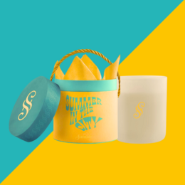 Tall, round, white scented candle with a teal-colored lid and handle. The candle has the text "Summer in the city" printed on the side in a yellow font.