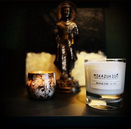 Tall, round, scented candle with a golden label and a decorative figurine in the background.