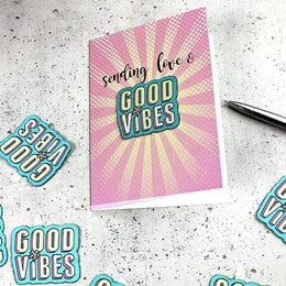 A greeting card with a pink and yellow background featuring the text "Sending love & good vibes" in a retro-style font. The image also includes a sticker-patch design with the text "GOOD VIBES" in a colorful, embroidered style.