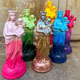Colorful tie-dye statues of the Virgin Mary, Notre-Dame de la Garde, in various vibrant hues including pink, green, gold, and blue. The statues feature the Virgin Mary holding the baby Jesus.