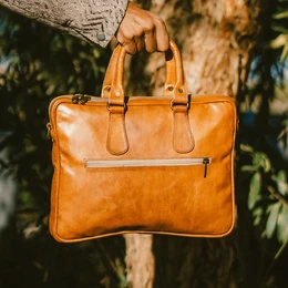  Handcrafted leather tech satchel and document case in a warm, caramel-colored leather with dual handles and a zippered compartment.