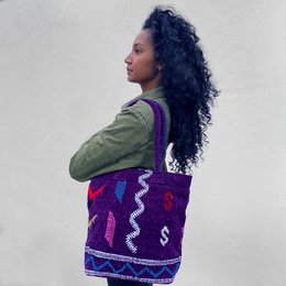  Venture Berber Rug Bag, a shoulder bag with a colorful, patterned design featuring a mix of blue, yellow, and red tones. The bag has a structured shape and appears to be made of a woven or textured material, likely inspired by traditional Berber rugs.