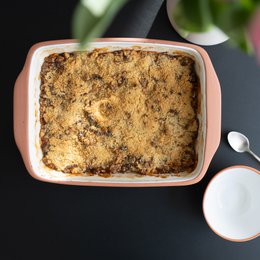 Rectangular ceramic baking dish with a golden brown crumble topping, likely containing a savory or sweet filling. Bright green lime or tomatillo halves arranged around the dish.