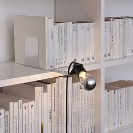 A modern, minimalist wall light with a sleek, cylindrical design and a single bulb. The light is mounted on a white shelf with books, creating a stylish and functional lighting solution for a home or office space.