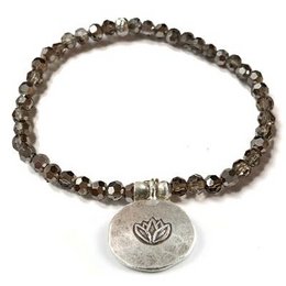 "Lotus bracelet - smoke: A delicate charm bracelet featuring a lotus flower charm and strands of smoky quartz beads. The bracelet has a natural, earthy style that would complement a variety of outfits."

The product is a charm bracelet featuring a lotus flower charm and strands of smoky quartz beads. The bracelet has a natural, earthy style with a neutral color palette of grays and browns. The materials used are likely semi-precious stones or glass beads. The overall shape of the bracelet is delicate and feminine, with the lotus charm adding a symbolic touch.