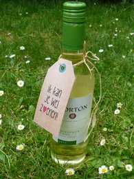 A bottle of white wine with a green label and a gift tag with the text "Vin je lief" (Dutch for "Love the wine"). The image shows the wine bottle in a grassy setting with white flowers.