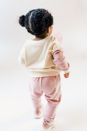 Sustainable high-waisted baby trousers in mauve velour, made with 100% GOTS certified organic cotton. Available in sizes 56, 62, 68, 74, 80, 86, and 92.
The image shows a young child wearing a gray top and pink bottoms. The child has curly dark hair and is sitting on a pink chair. The product appears to be a high-waisted pair of baby trousers in a mauve velour material, made from 100% GOTS certified organic cotton.