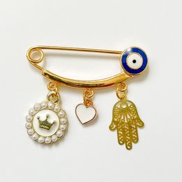 A gold-colored pin with a dark blue evil eye charm, a heart-shaped charm, and a hamsa hand charm. The pin is designed as a lucky charm for babies and can be used as a gift for birth.