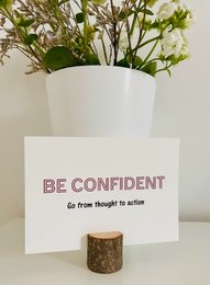 A6 greeting card with the text "Be confident. Go from thought to action" printed in red on a cream-colored background. The card is displayed in a white vase with greenery.