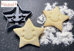 Smiling star-shaped baking decoration, made of light-colored dough, with a black star-shaped cookie cutter visible in the background.