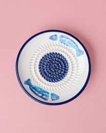A round, shallow ceramic dish with a blue and white pattern featuring a central spiral design. The dish appears to be used for grating or shredding food items, as indicated by the hand holding a tomato and grating it in the second image.