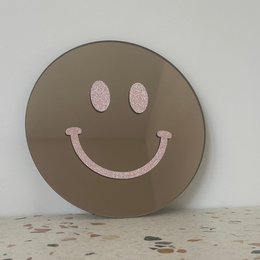 A round, mirrored wall decor with a pink glittery smiley face design.