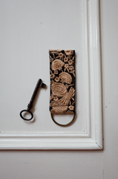 Decorative keychain with intricate floral pattern in brown and beige tones, featuring a D-ring and musketon for attachment.